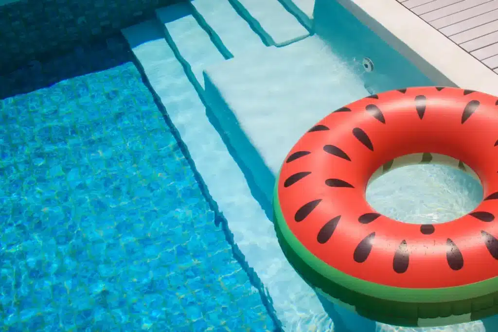 Bouée pastèque et piscine bleue pour un été relaxant Bouée pastèque flottant sur l'eau bleue d'une piscine ensoleillée avec des marches immergées.