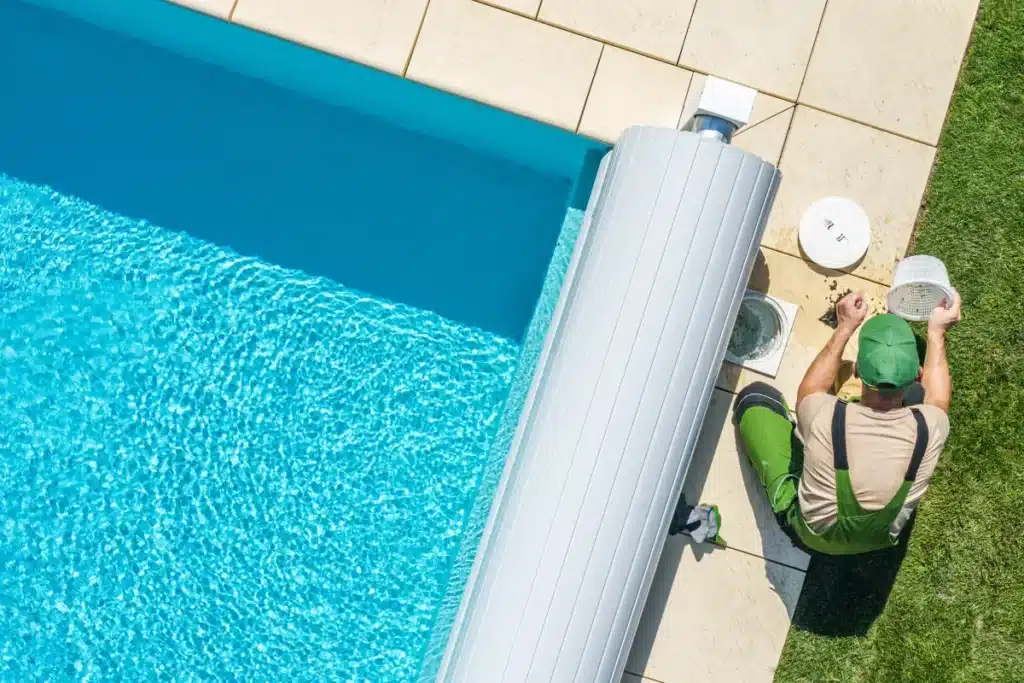 Entretien de piscine : nettoyage du skimmer par un pro Technicien en salopette verte entretenant le skimmer d'une piscine avec volet roulant.