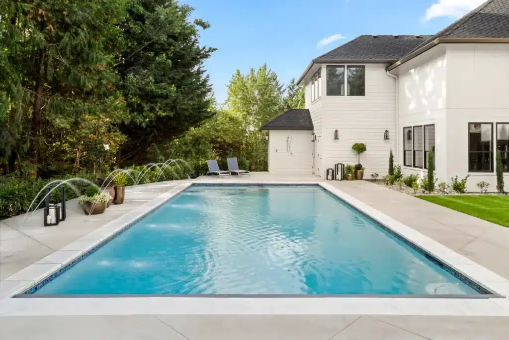 Piscine moderne avec jets d'eau et jardin luxueux Piscine rectangulaire moderne avec jets d'eau, terrasse en pierre et jardin luxuriant devant une maison blanche.