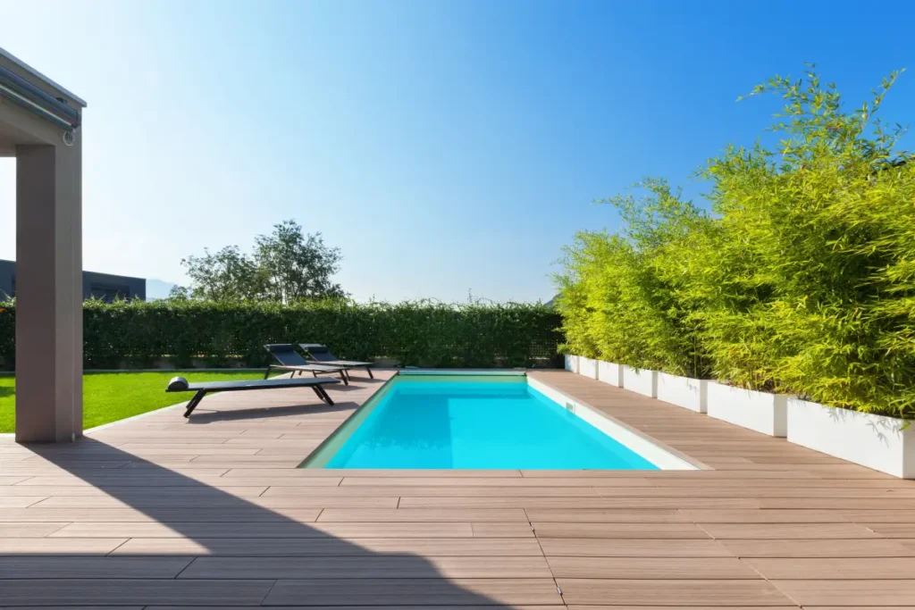 Piscine moderne avec terrasse en bois et bambous décoratifs Piscine rectangulaire avec terrasse en bois, transats noirs et haie de bambous sous un ciel bleu dégagé.