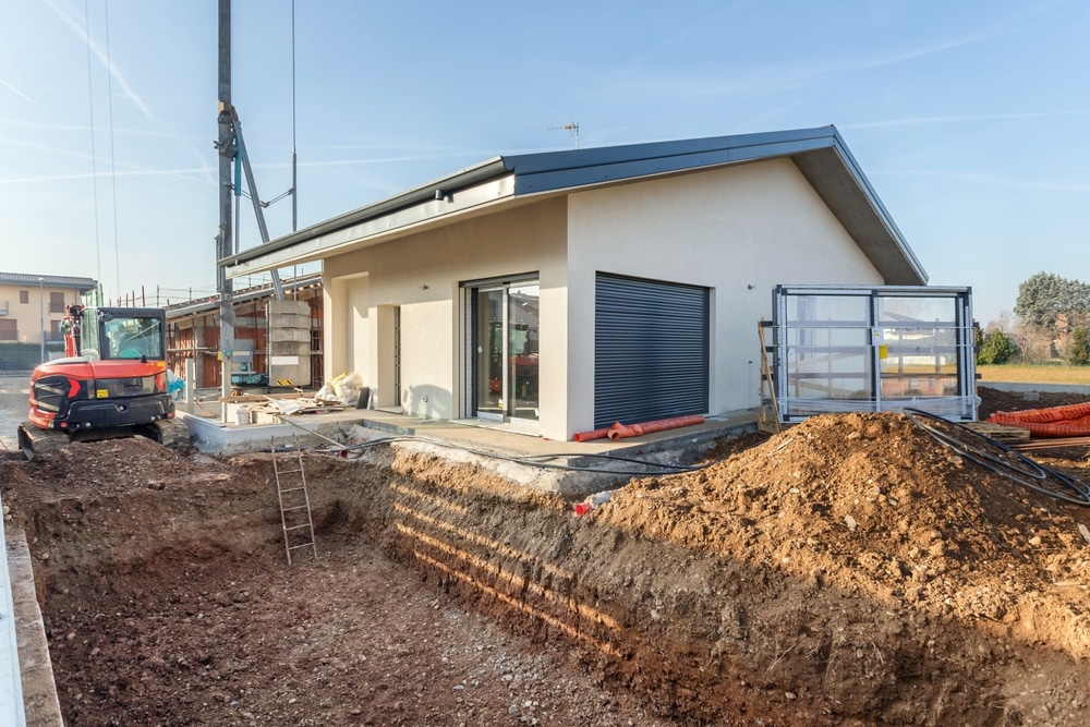 Construction maison moderne : excavation et terrassement Chantier de maison moderne avec excavation pour piscine et pelle mécanique orange sous un ciel bleu.