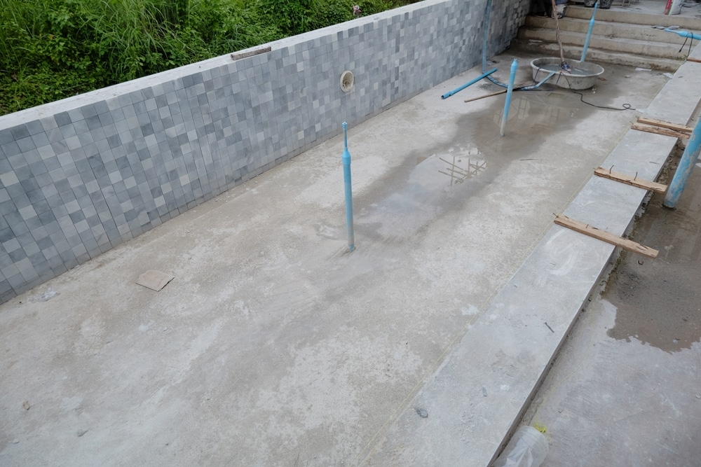 Construction de piscine carrelée : étapes et gros œuvre Piscine en construction avec parois carrelées grises, fond en béton brut et tuyaux PVC sur un chantier extérieur.