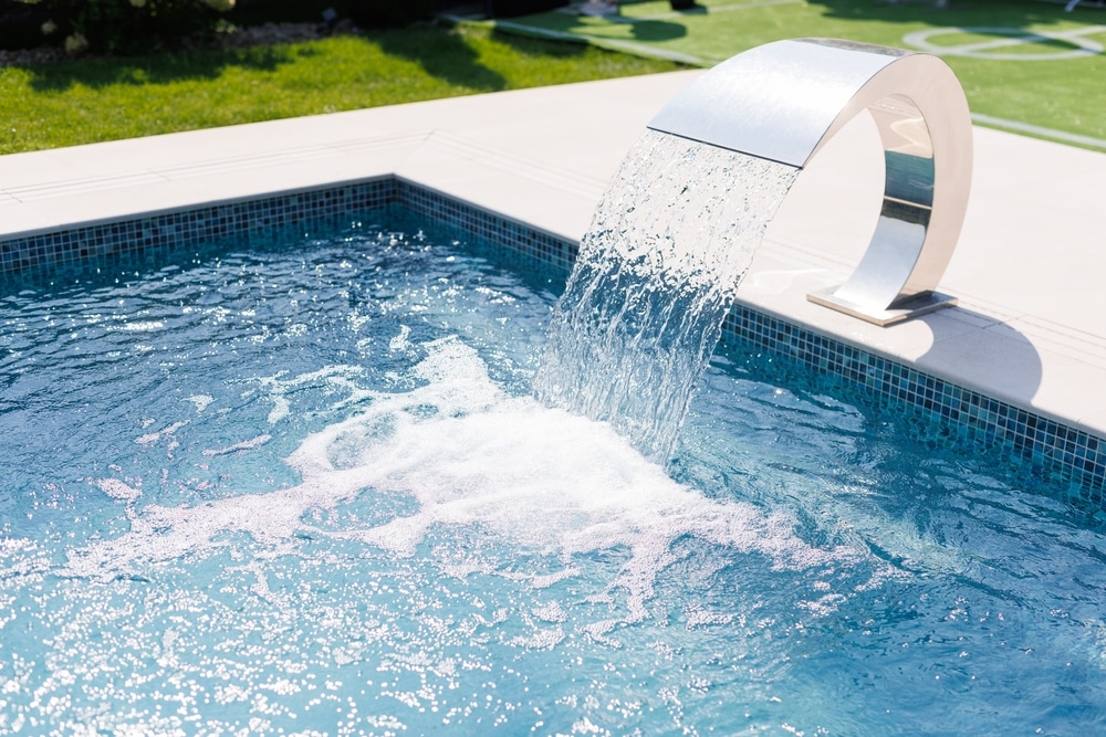 Fontaine de piscine cascade en inox design et moderne Cascade de piscine en inox design versant de l'eau cristalline dans un bassin carrelé bleu sous le soleil.