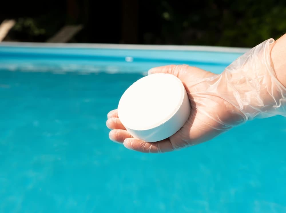 Galet de chlore piscine : entretien et traitement de l'eau Main gantée tenant un gros galet de chlore blanc au-dessus d'une piscine bleue pour l'entretien et le traitement de l'eau.