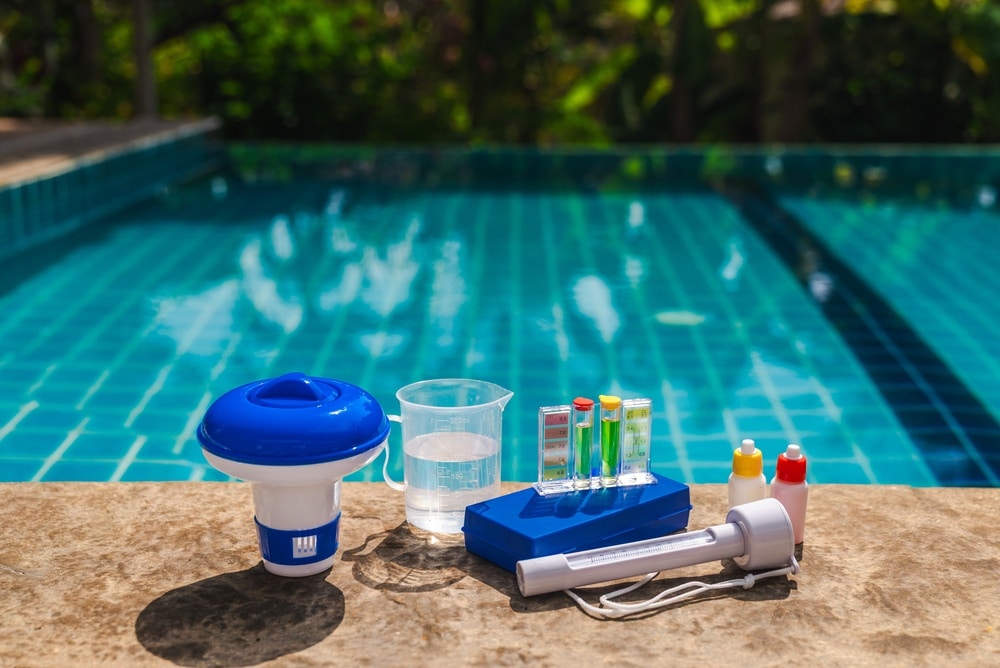 Entretien piscine : kit de test pH et doseur de chlore Kit d'entretien de piscine avec doseur de chlore, testeur pH et thermomètre au bord d'un bassin bleu ensoleillé.