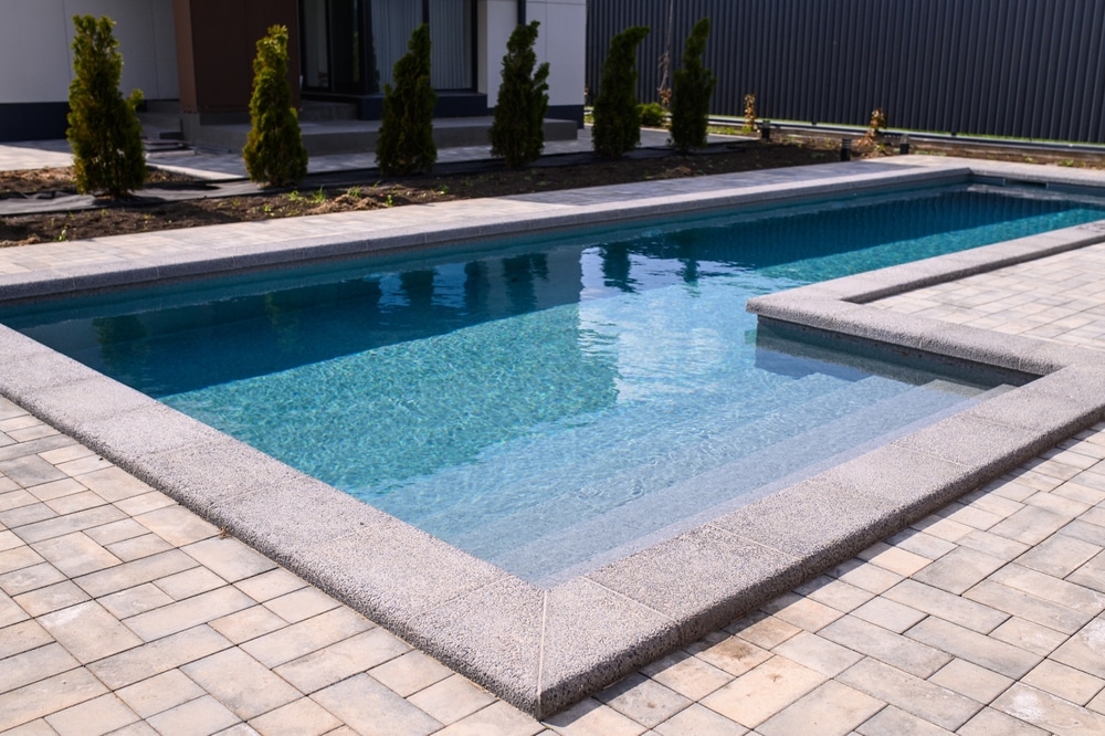 Piscine en L moderne avec margelles et terrasse pavée Piscine en L moderne aux eaux cristallines avec margelles grises et terrasse pavée dans un jardin paysager.