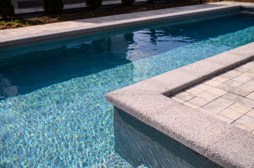 Aménagement de Piscine Moderne avec Margelles en Pierre Piscine moderne avec margelles en pierre grise, eau turquoise scintillante et terrasse en pavés beiges.