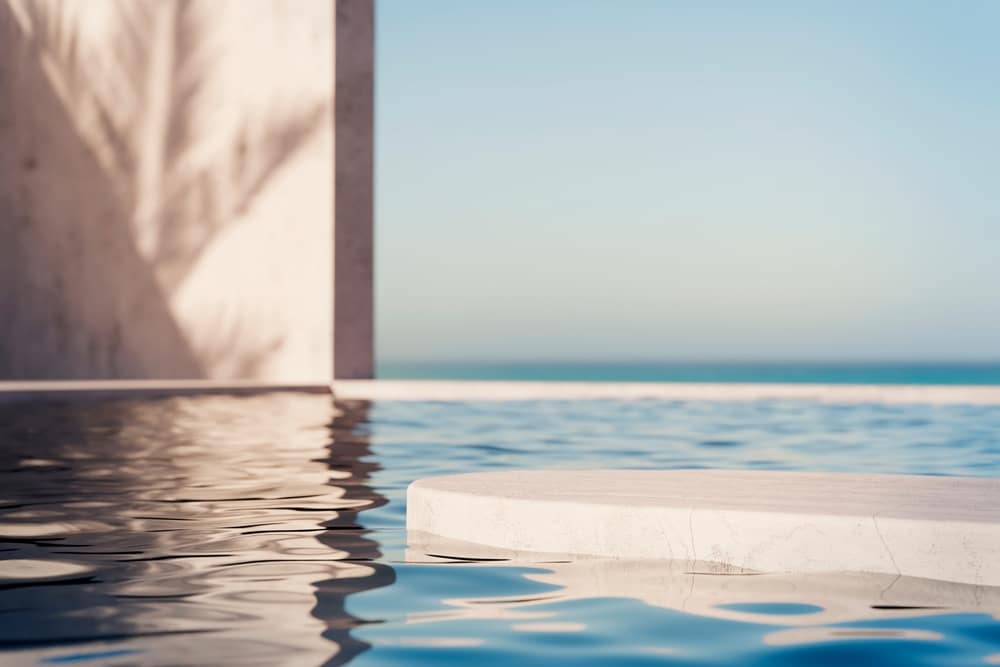 Podium minimaliste sur l'eau pour présentation produit Podium en pierre blanche sur une eau bleue calme avec ombre de palmier et vue sur la mer à l'horizon.