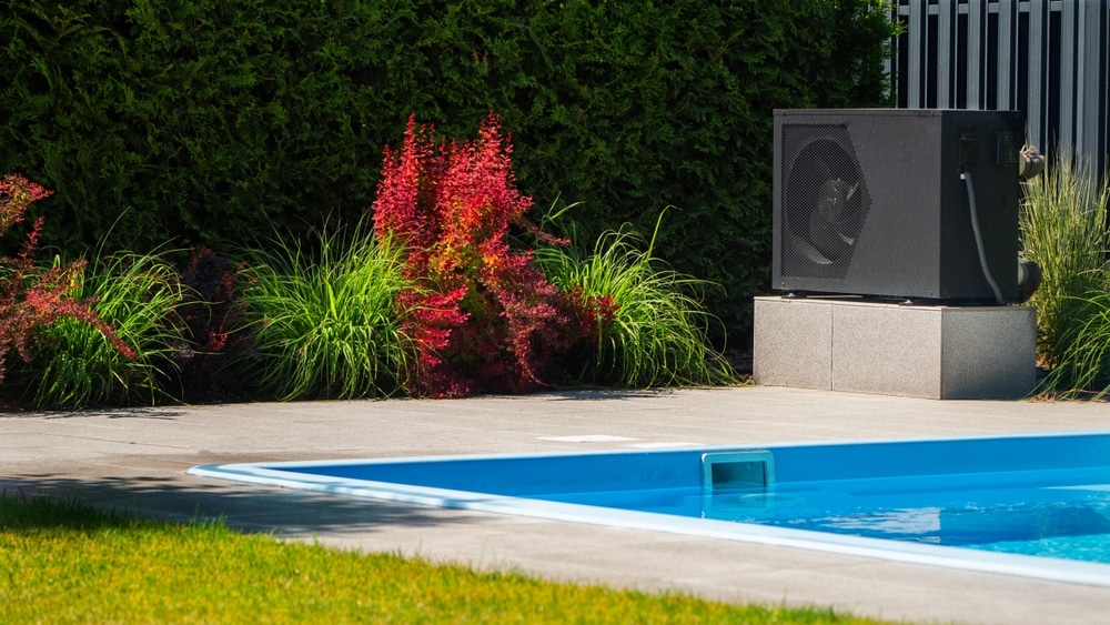 Pompe à chaleur pour piscine : confort thermique extérieur Pompe à chaleur noire installée sur une dalle près d'une piscine bleue avec un jardin paysager en arrière-plan.